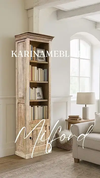 Prezentacja wideo: Wysoka, wąska biblioteczka z jasnego, postarzanego drewna mango (model Milford) z otwartymi półkami i dolną szafką, wypełniona książkami i zdjęciami w ramkach, ustawiona w jasnym, przytulnym wnętrzu na tle ściany z białą boazerią, obok jasnej sofy i drewnianego stolika z lampką, w pobliżu okna z łukiem – Meble Rustykalne