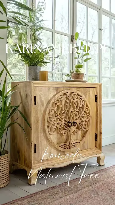 Prezentacja wideo: Niska dwudrzwiowa komoda z jasnego, naturalnego drewna mango (model Natural Tree) z frontami ozdobionymi dużym, rzeźbionym motywem drzewa życia wpisanego w okrąg, wyposażona w małe, ciemne uchwyty i osadzona na dekoracyjnych, giętych nóżkach, udekorowana zielonymi roślinami doniczkowymi oraz karafką na blacie, zaprezentowana w jasnej oranżerii przy dużych oknach ze szprosami, stojąca na szarej podłodze obok klasycznego dywanu – Meble Indyjskie / Boho