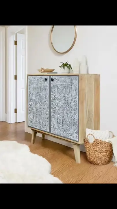 Prezentacja wideo: Dwudrzwiowa komoda z jasnego drewna mango (model Lumi) z frontami ozdobionymi misternymi, szaro-białymi rzeźbieniami, osadzona na skośnych nóżkach w stylu retro, udekorowana małą rośliną doniczkową, wazami oraz drewnianą miseczką, zaprezentowana w jasnym wnętrzu pod okrągłym lustrem, stojąca na drewnianej podłodze obok puszystego, białego dywanu i plecionego kosza – Meble Boho