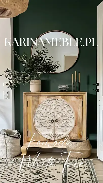 Prezentacja wideo: Drewniana komoda z jasnego drewna mango (model White Tree) z dużym, okrągłym, bielonym i bogato rzeźbionym motywem drzewa życia na drzwiczkach, ustawiona na tle eleganckiej, ciemnozielonej ściany z okrągłym lustrem, udekorowana gałązkami w wazonie i złotymi świecznikami, na tle wzorzystego dywanu i plecionego kosza – Meble Orientalne