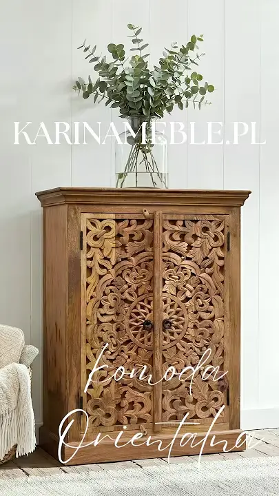 Prezentacja wideo: Wysoka dwudrzwiowa komoda z drewna mango w kolorze jasnego orzecha z frontami ozdobionymi kunsztownym, rzeźbionym motywem roślinnym z centralną rozetą i mandalą, wyposażona w małe okrągłe uchwyty, udekorowana dużym szklanym wazonem z zielonymi gałązkami eukaliptusa, zaprezentowana w jasnym, przytulnym wnętrzu na tle białej ściany z pionowych paneli, stojąca na drewnianej podłodze przy jasnym dywanie obok fragmentu fotela z kocem z frędzlami – Meble Orientalne / Indyjskie