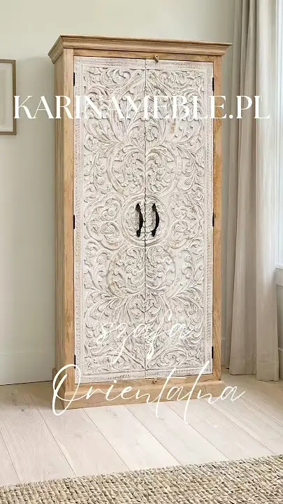 Prezentacja wideo: Wysoka dwudrzwiowa szafa o wysokości 200 cm z drewna mango z korpusem w naturalnym odcieniu oraz białymi, przecieranymi frontami ozdobionymi misternie rzeźbionym motywem roślinnym w stylu shabby chic, wyposażona w ciemne, metalowe uchwyty, zaprezentowana w jasnym wnętrzu na jasnej drewnianej podłodze obok strukturalnego, plecionego dywanu – Meble Indyjskie / Shabby Chic